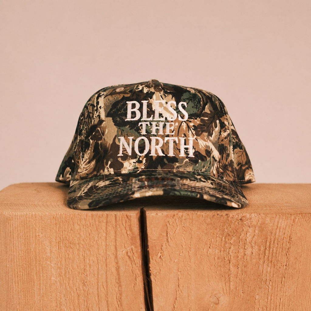 A camouflage trucker cap with the words 'BLESS THE NORTH' printed in white on the front, resting on a wooden block against a solid beige background.