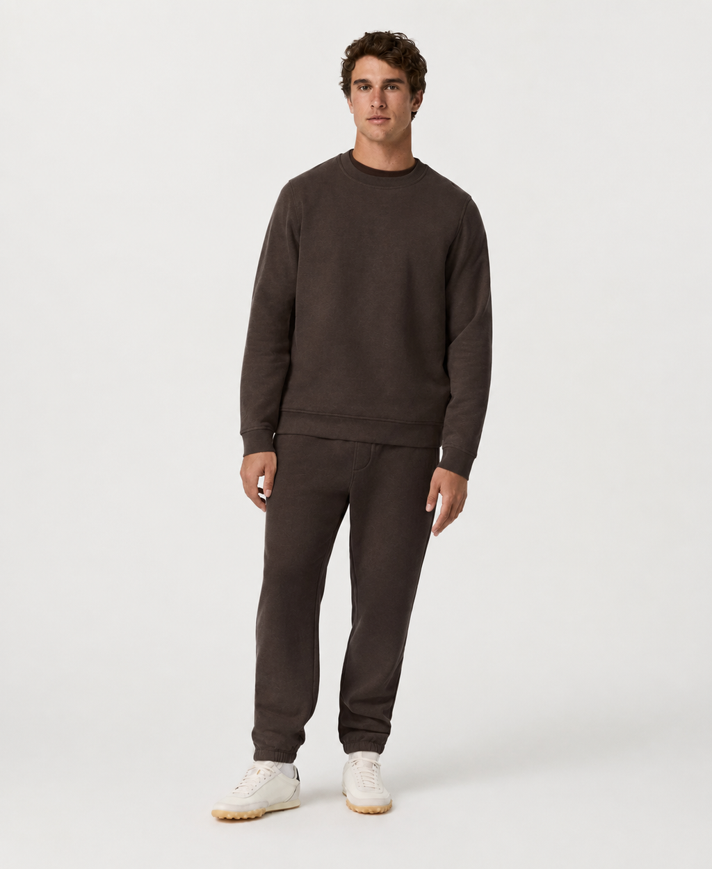 A man modeling a dark brown crewneck sweatshirt and matching sweatpants set, paired with white sneakers, against a plain light gray studio background.