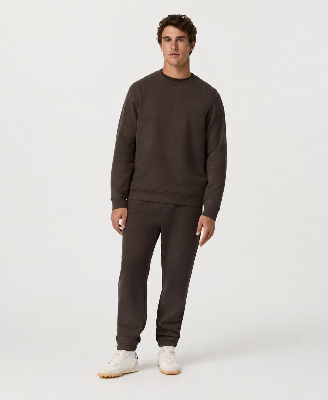 A man modeling a dark brown crewneck sweatshirt and matching sweatpants set, paired with white sneakers, against a plain light gray studio background.