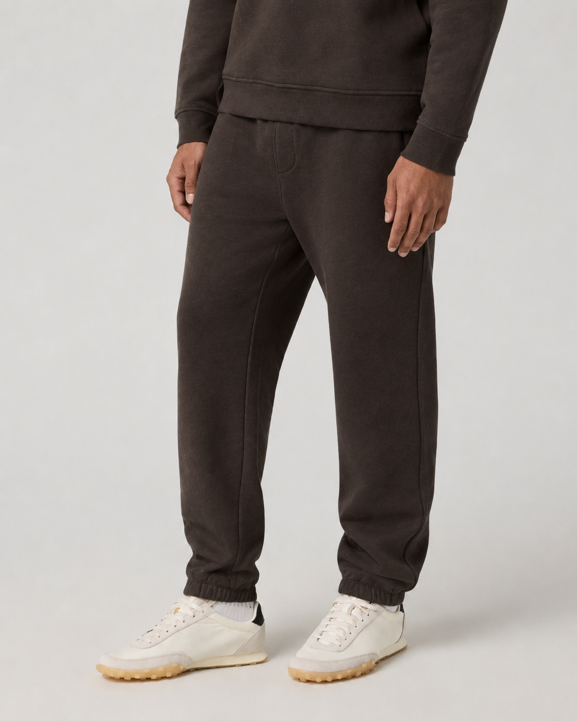 A person wearing dark brown jogger sweatpants with an elastic waistband and cuffs, paired with white sneakers, shown from the waist down on a light gray studio background.