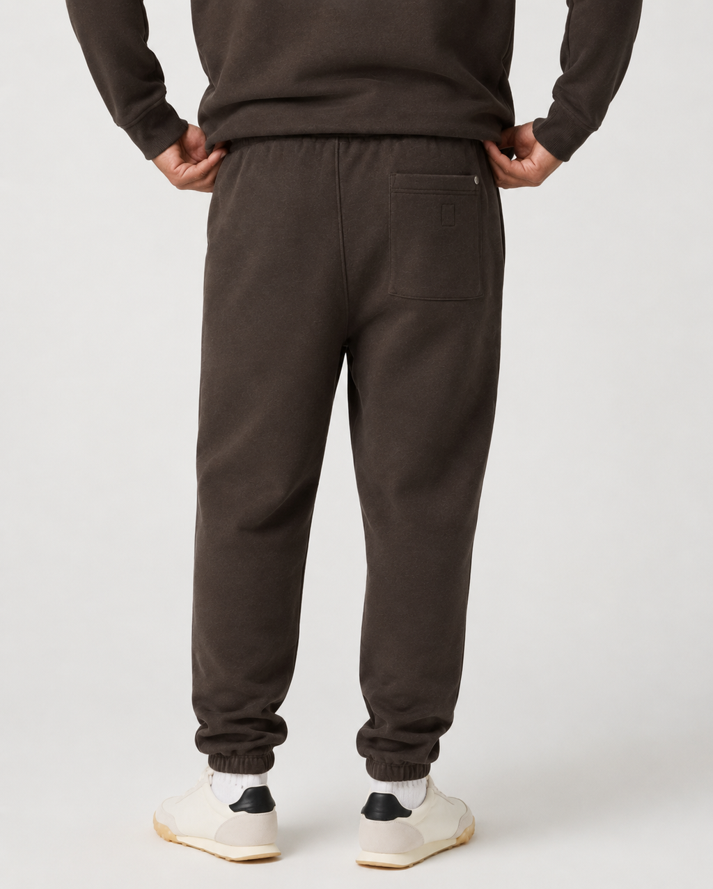 Rear view of a person wearing dark brown sweatpants with an elastic waistband, back pocket, and ribbed ankle cuffs, paired with white sneakers against a light gray studio background.
