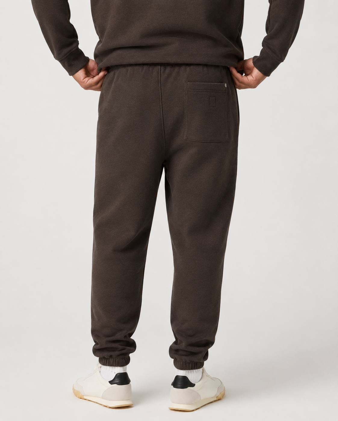 Rear view of a person wearing dark brown sweatpants with an elastic waistband, back pocket, and ribbed ankle cuffs, paired with white sneakers against a light gray studio background.