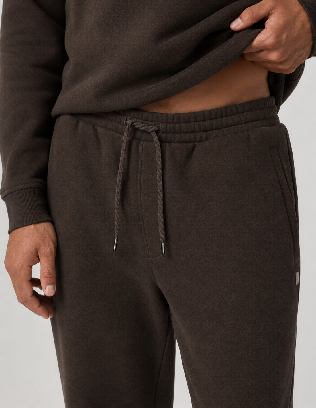 Close-up of a person wearing dark brown fleece sweatpants with a matching drawstring waist and a dark brown sweatshirt, shown against a light gray studio background.