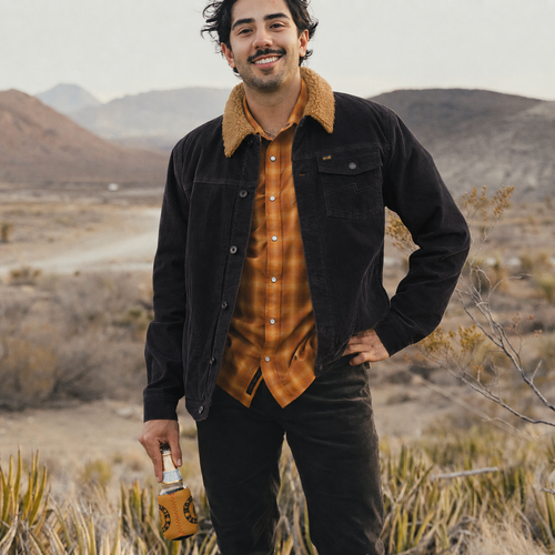A man standing in a desert landscape wearing a black corduroy jacket with a tan sherpa collar over an orange-brown plaid button-up shirt and black pants, holding an orange tumbler.