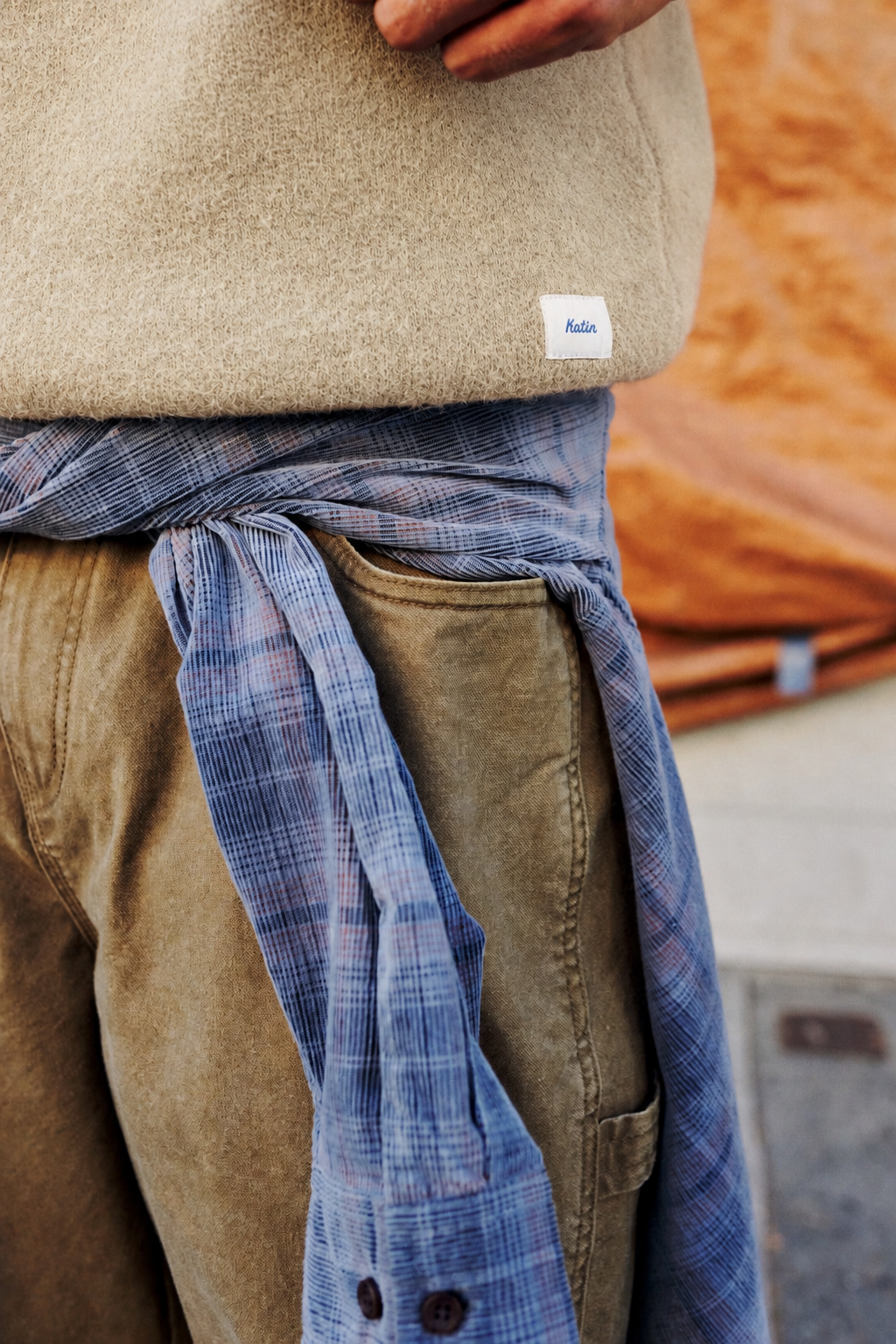 Close-up of a person wearing a beige knit sweater, brown pants, and a blue plaid button-up shirt tied around the waist. A small white tag on the sweater reads Audie.