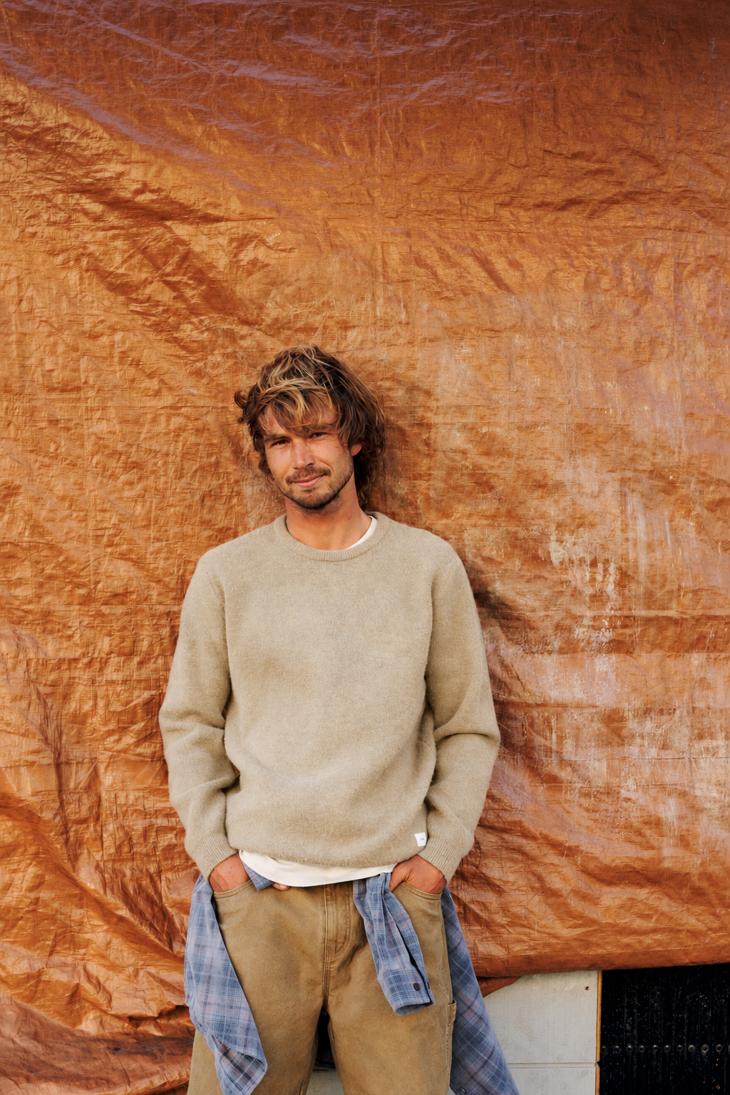 A man with messy light brown hair stands against a wrinkled orange-brown backdrop, wearing a beige crewneck sweater over a white undershirt, tan pants, and a blue plaid shirt tied around his waist.