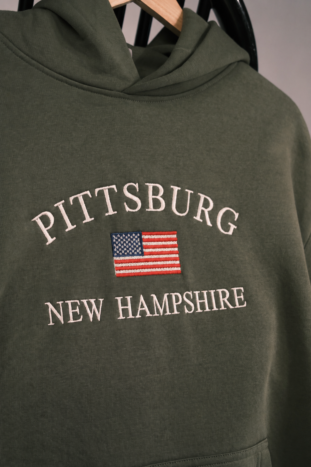 An olive green pullover hoodie hanging on a wooden hanger, featuring 'PITTSBURG' and 'NEW HAMPSHIRE' in cream arched text with an embroidered American flag in the center.