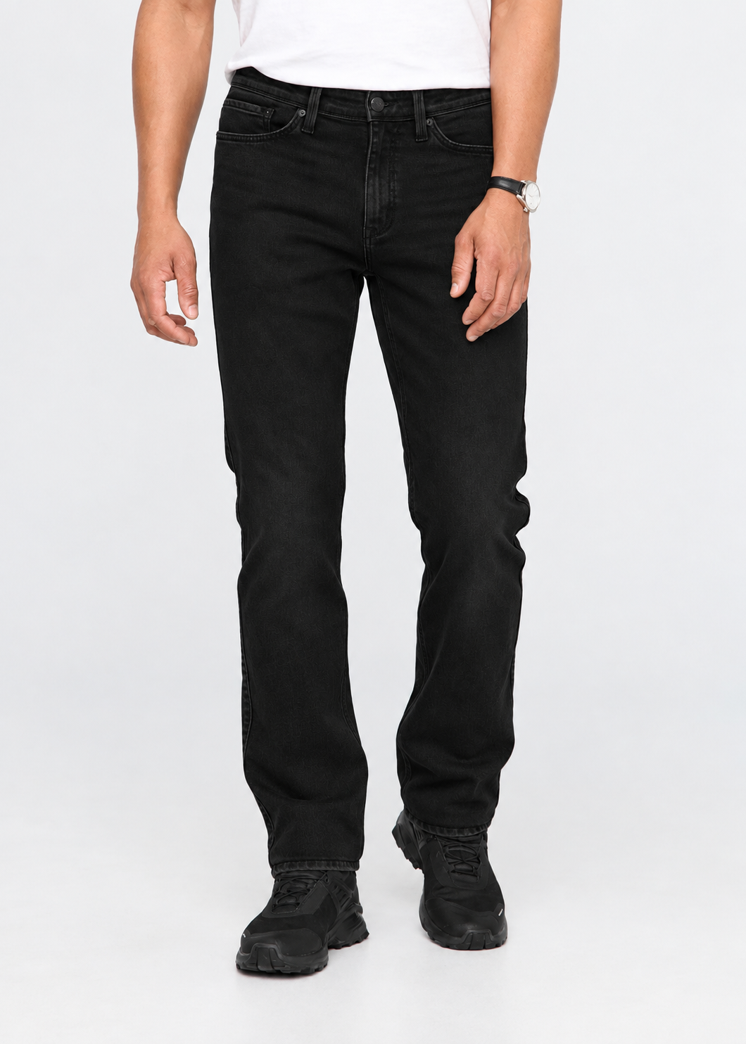 A person wearing black slim-fit jeans, a white t-shirt, and black sneakers, standing against a light gray studio background.