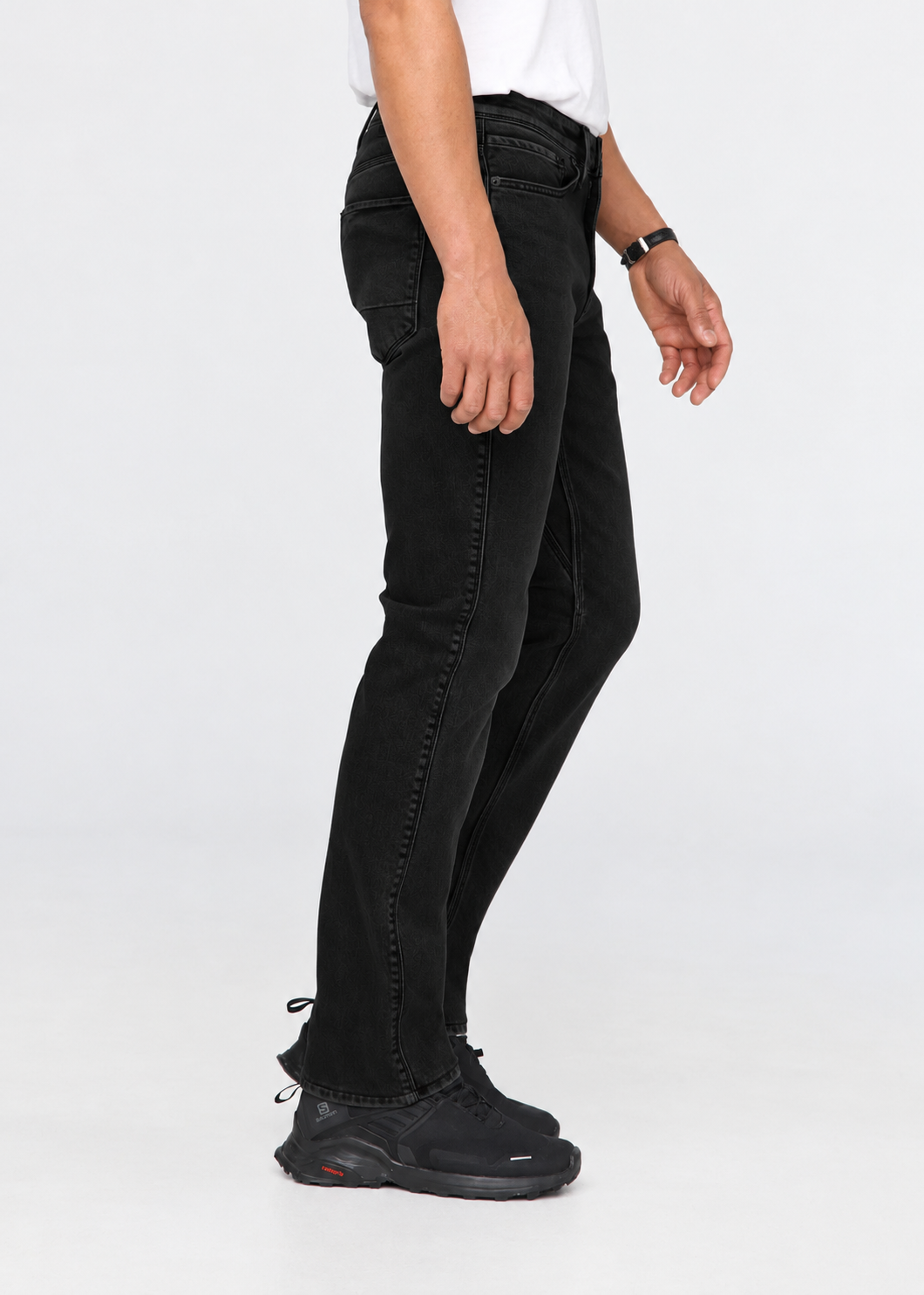 A side-profile view of a person wearing slim-fit black jeans, a white t-shirt, and black sneakers against a plain white studio background.