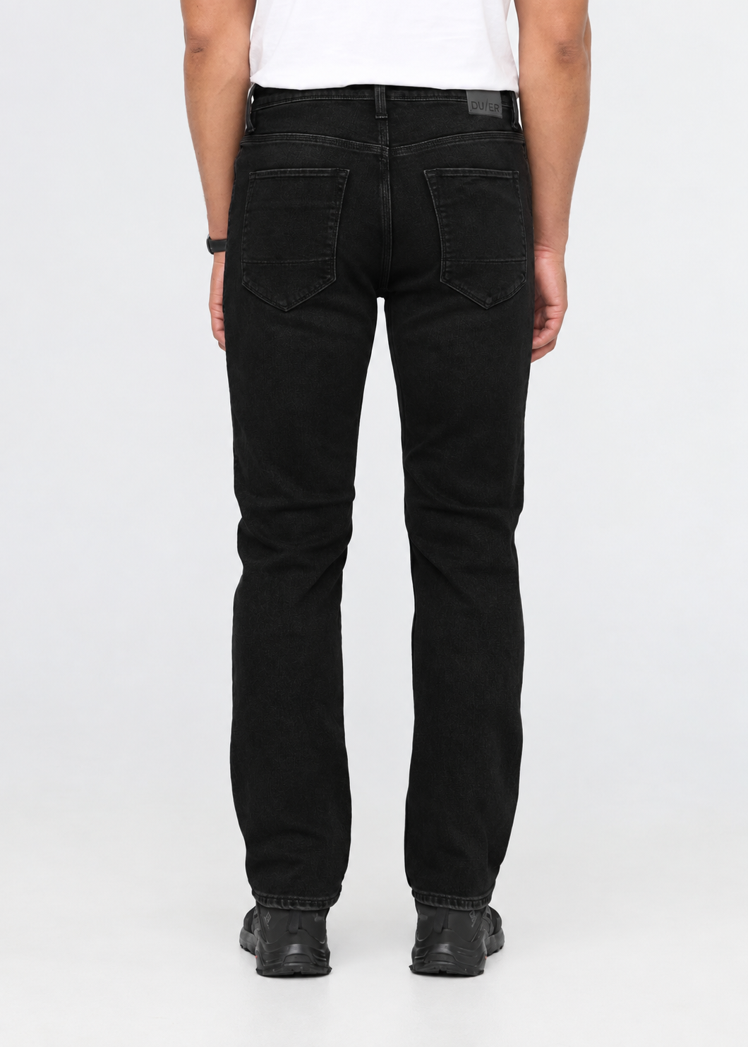 Rear view of a person wearing black jeans with a white shirt and black shoes, photographed against a light gray studio background.