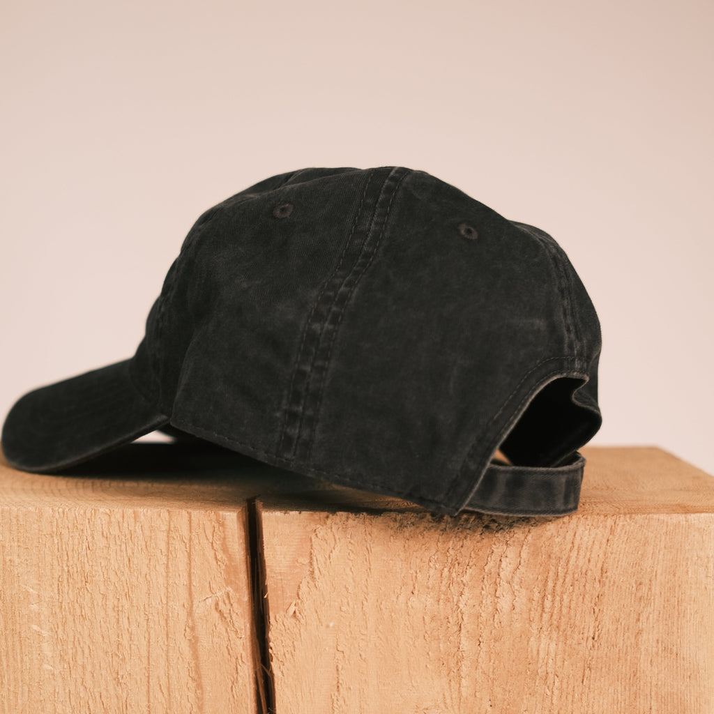 A black washed baseball cap resting on a light wood block against a solid beige background.