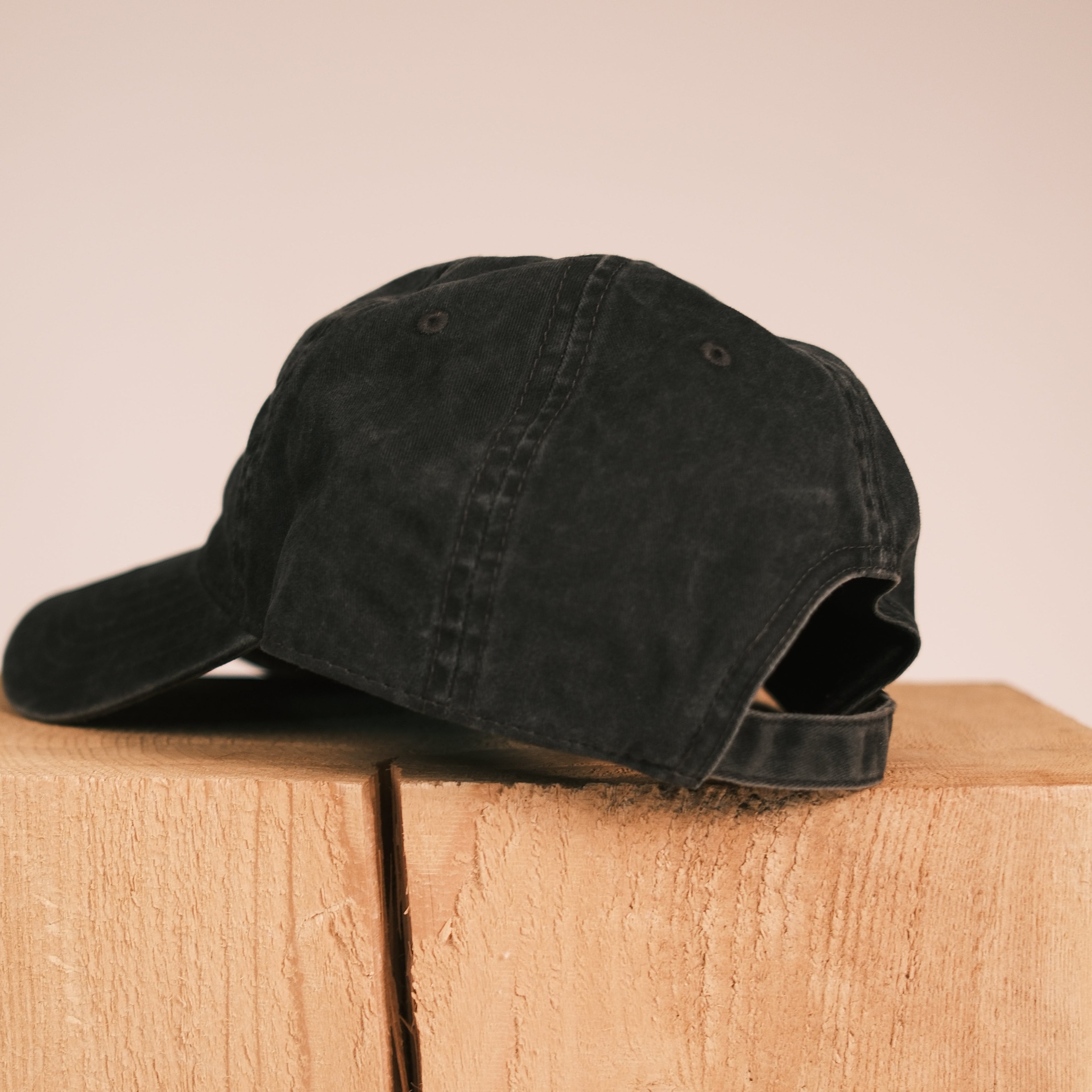 A black washed baseball cap resting on a light wood block against a solid beige background.