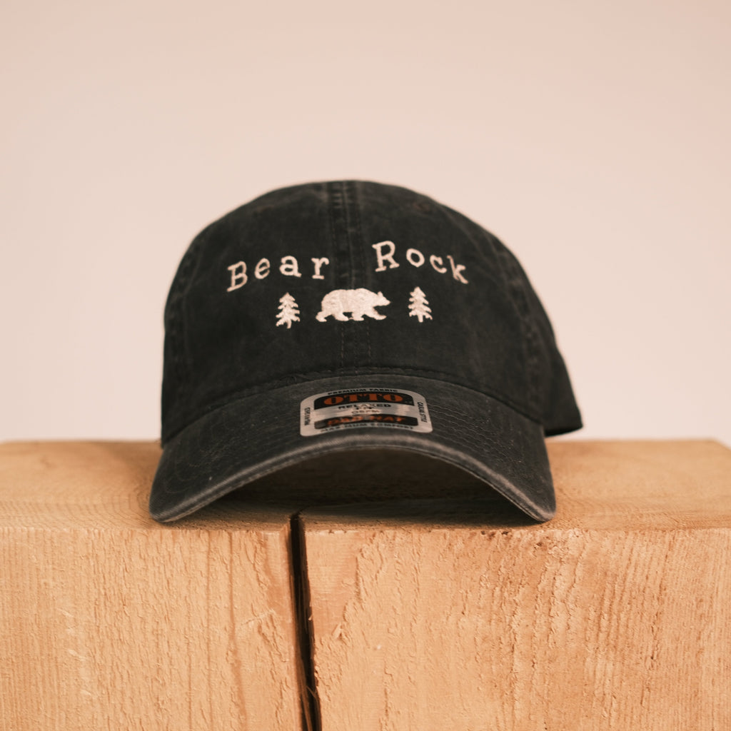 A black distressed baseball cap with 'Bear Rock' and a bear graphic embroidered on the front, displayed on a light wood block against a beige background.