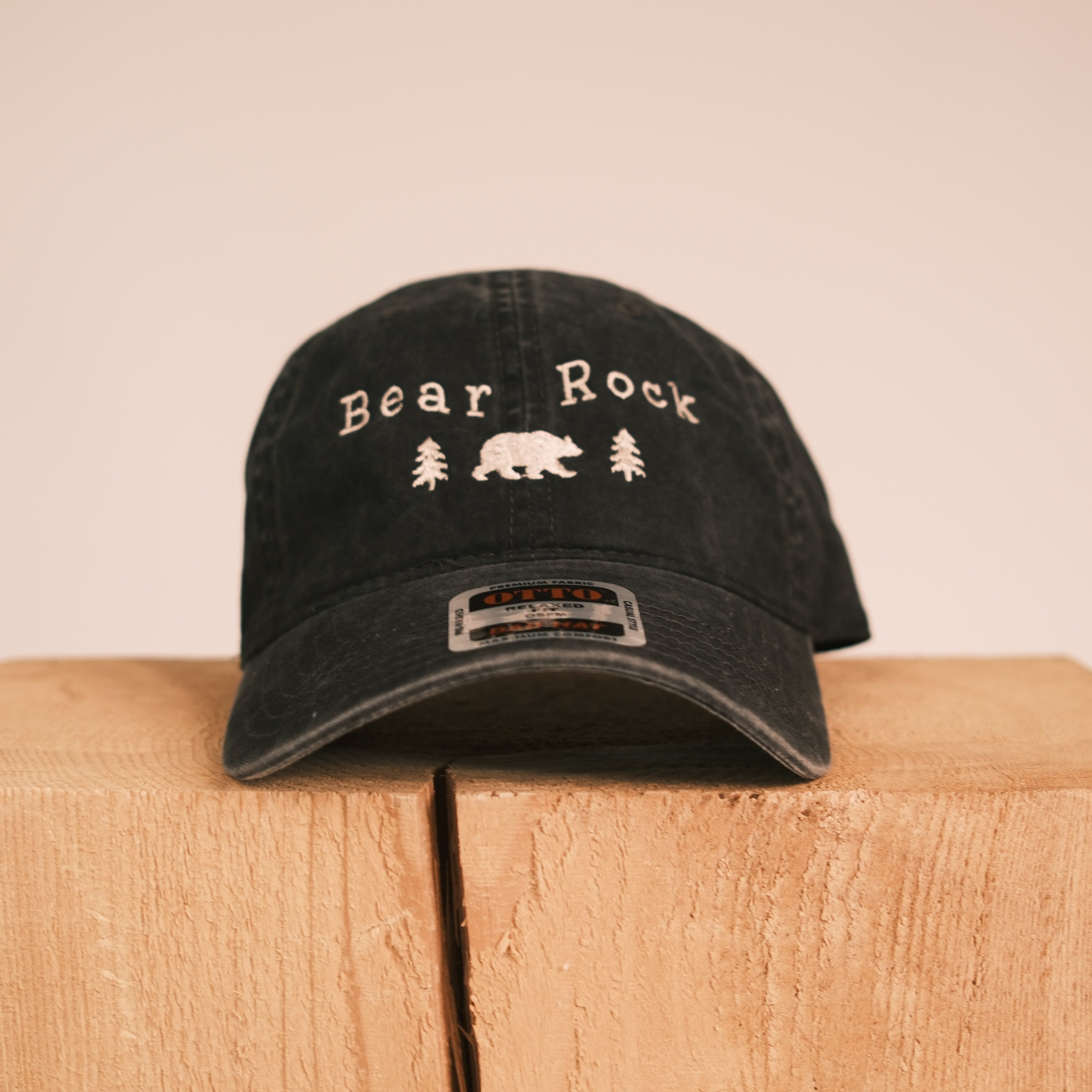 A black distressed baseball cap with 'Bear Rock' and a bear graphic embroidered on the front, displayed on a light wood block against a beige background.