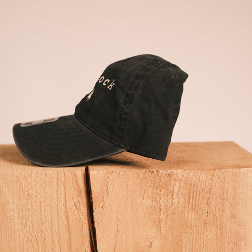 A black washed baseball cap with white embroidered text, resting on a light wood block against a solid beige background.
