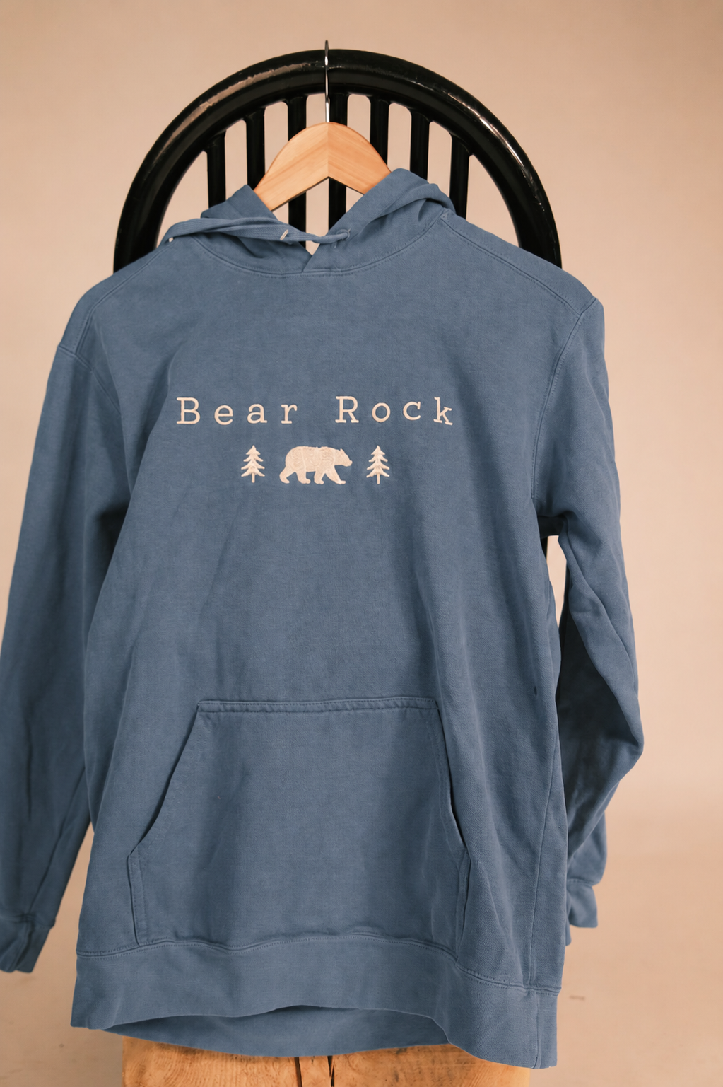 A dusty blue pullover hoodie with a white 'Bear Rock' graphic featuring a bear and pine trees, hanging on a wooden hanger against a black chair back.