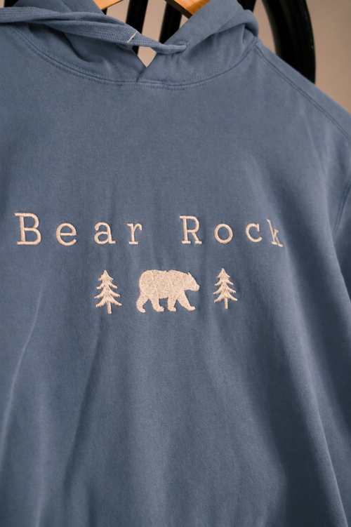 A close-up of a dusty slate blue hoodie featuring a cream-colored graphic on the chest with the text 'Bear Rock' and a central bear flanked by two pine trees.