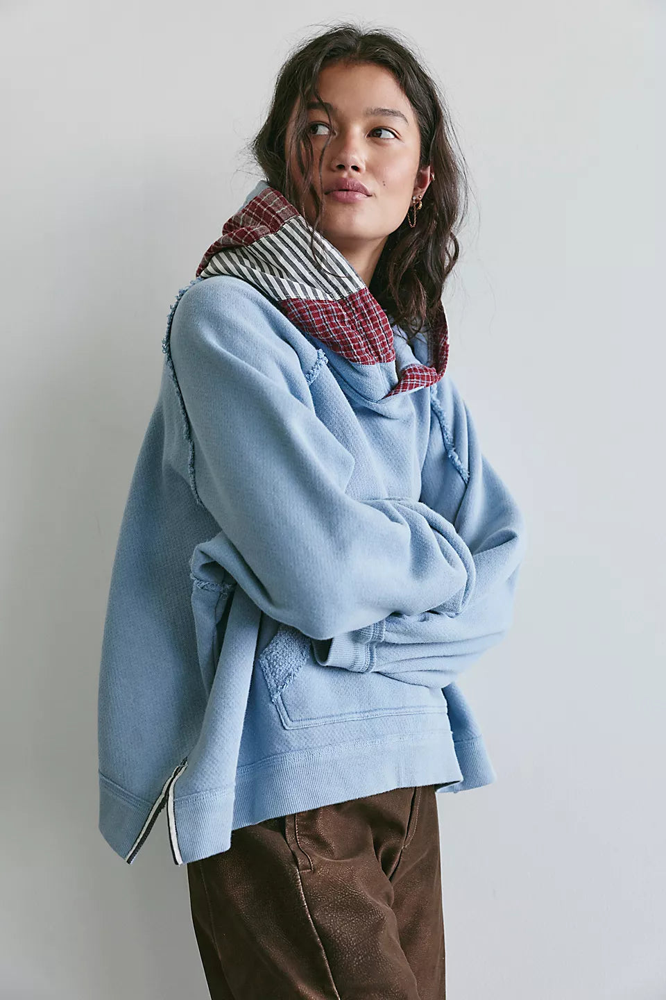 Woman with arms folded modeling light blue hooded sweatshirt with red plaid and black and white stripes on hood and brown pants