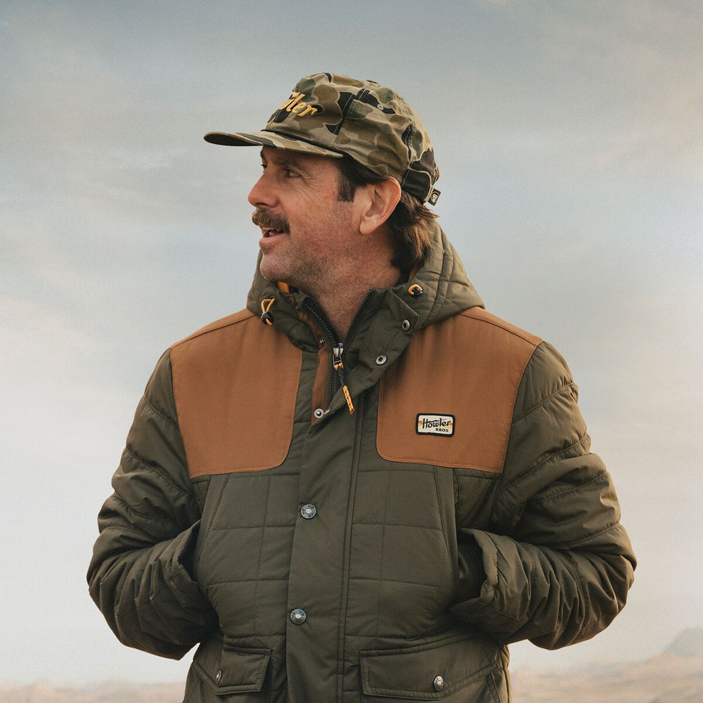 Man wearing a green and brown jacket with a camouflage cap, standing against a neutral background.