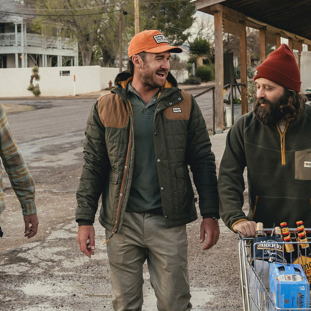 Two men in outdoor clothing standing on a street, one with a cart of beer.