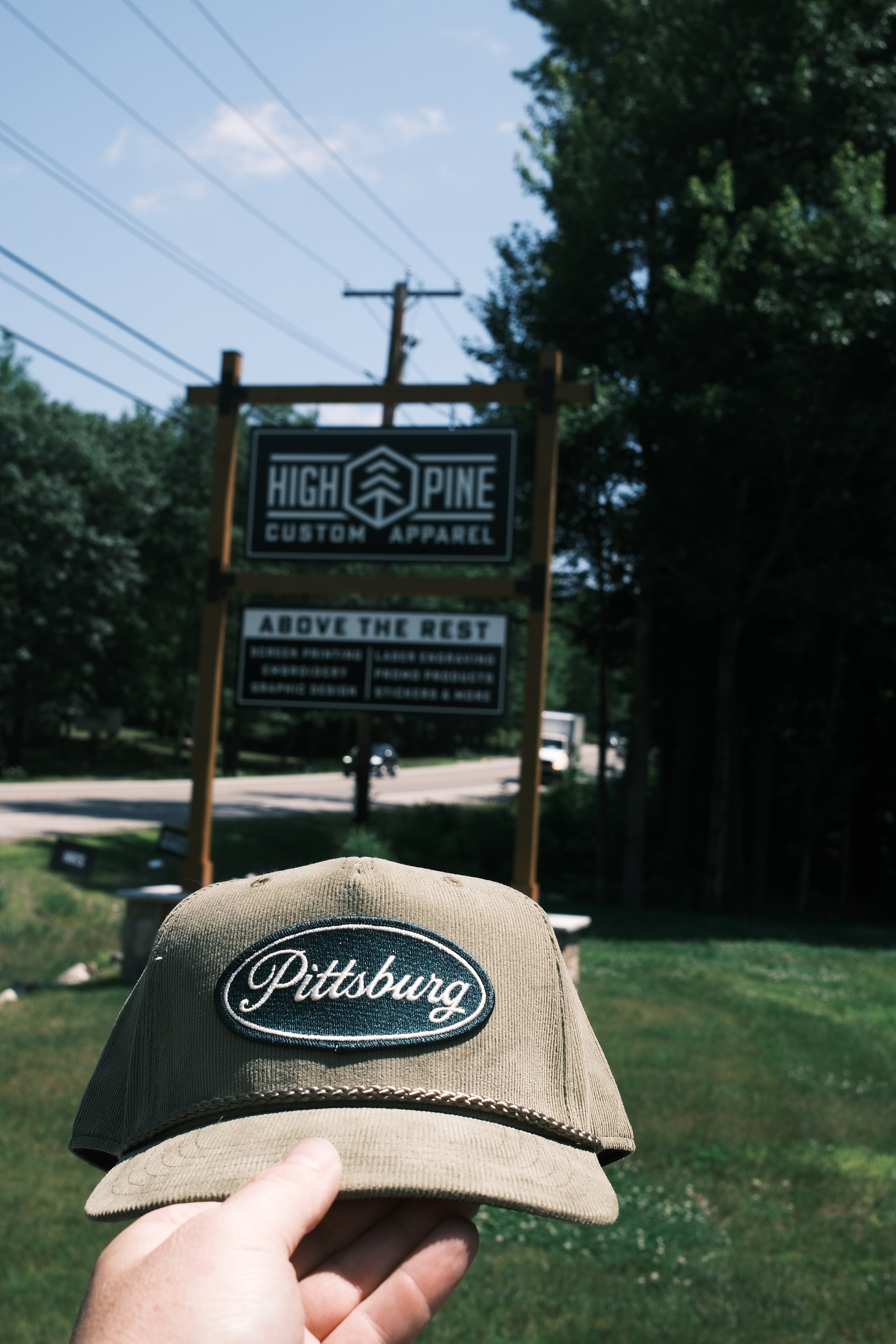 A hand holds a khaki baseball cap with a 'Pittsburg' embroidered patch in the foreground, with a wooden sign for 'High Pine Custom Apparel' and trees in the background.