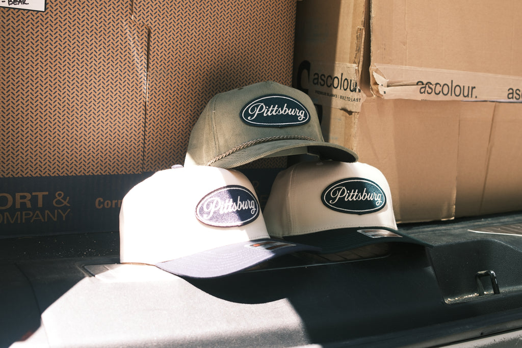 Three Pittsburgh baseball caps in different colors (olive green, cream, and dark gray) arranged on top of cardboard shipping boxes.