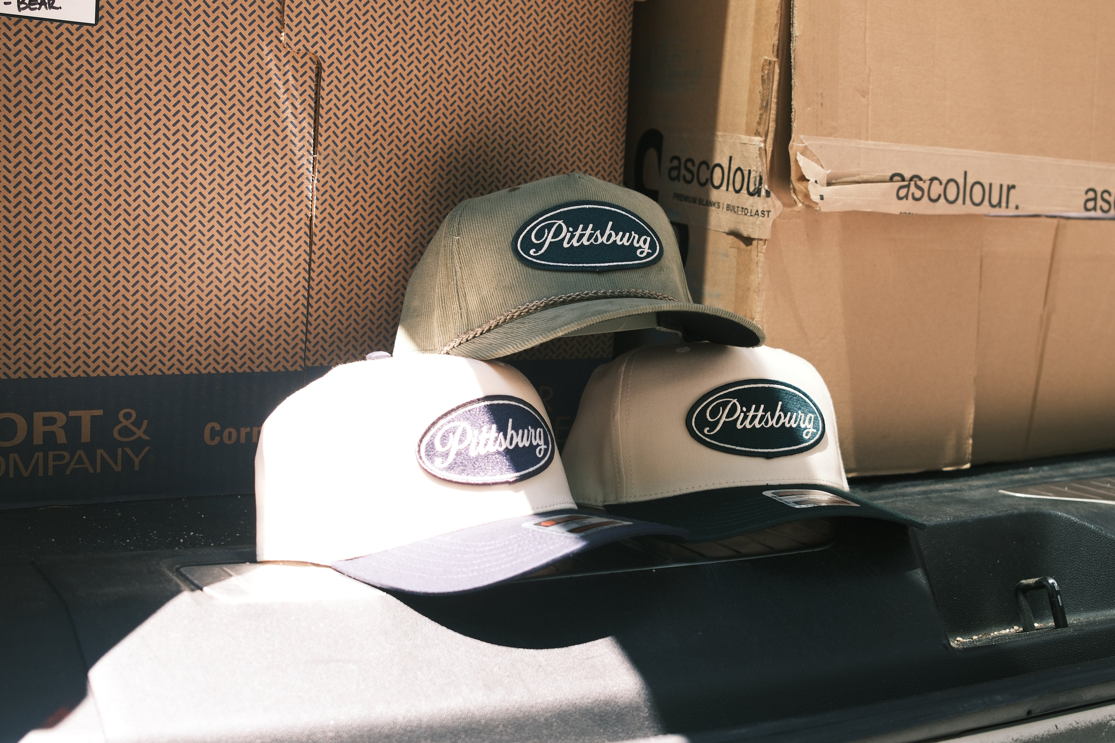 Three Pittsburgh baseball caps in different colors (olive green, cream, and dark gray) arranged on top of cardboard shipping boxes.
