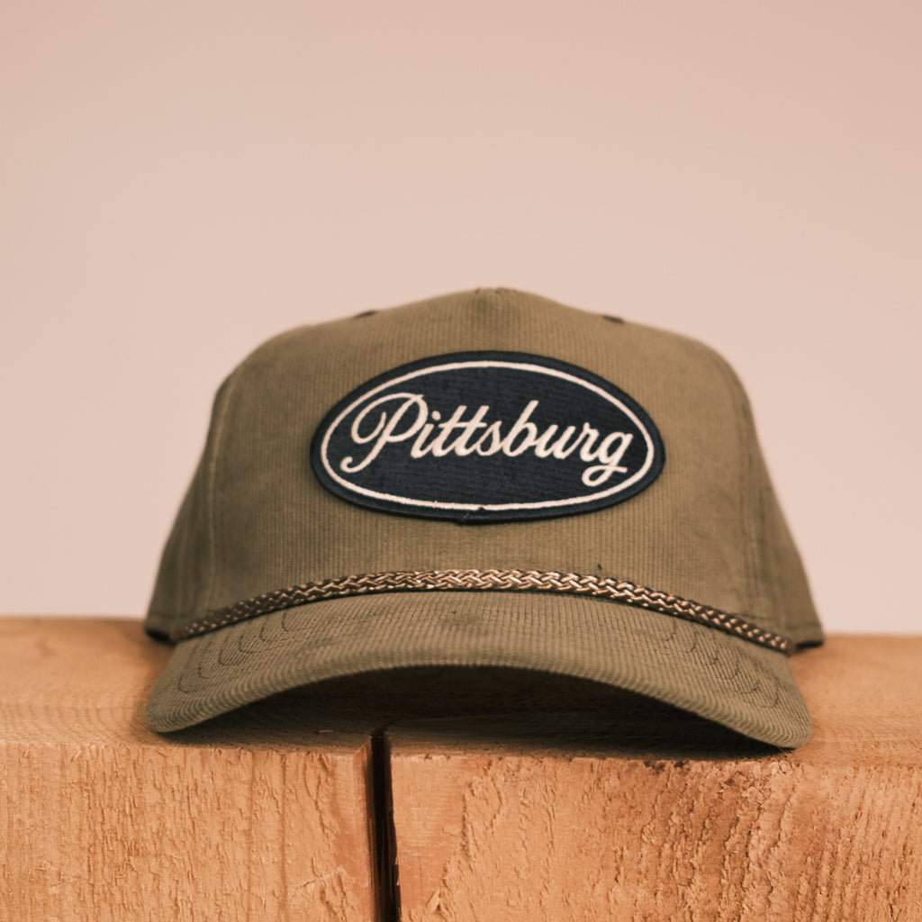 An olive green baseball cap with a black oval 'Pittsburg' embroidered patch on the front, resting on a rustic wooden block against a solid beige background.