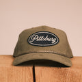 An olive green baseball cap with a black oval 'Pittsburg' embroidered patch on the front, resting on a rustic wooden block against a solid beige background.