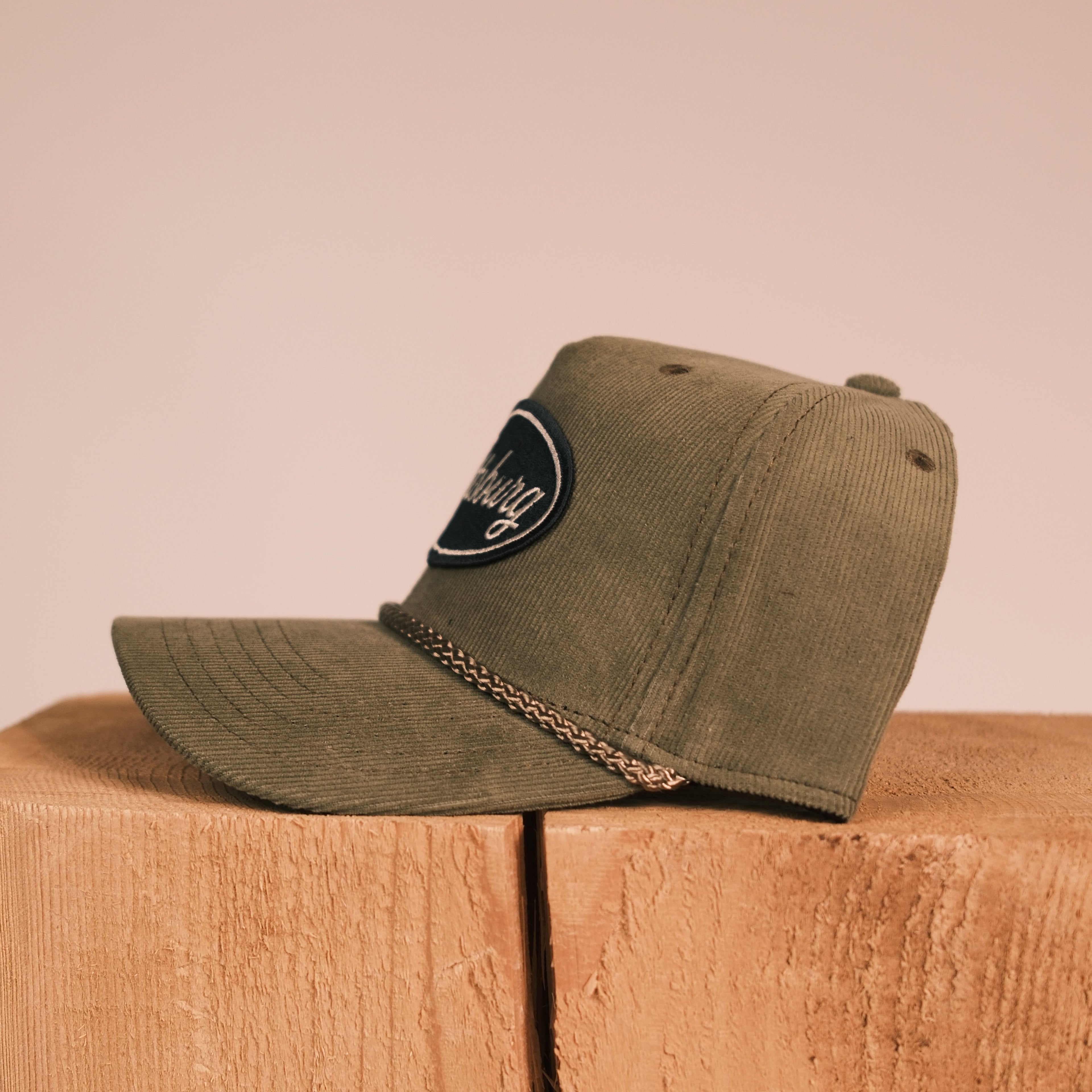 An olive green corduroy baseball cap with a black embroidered patch and a gold chain detail, resting on a rustic wooden block against a solid beige background.