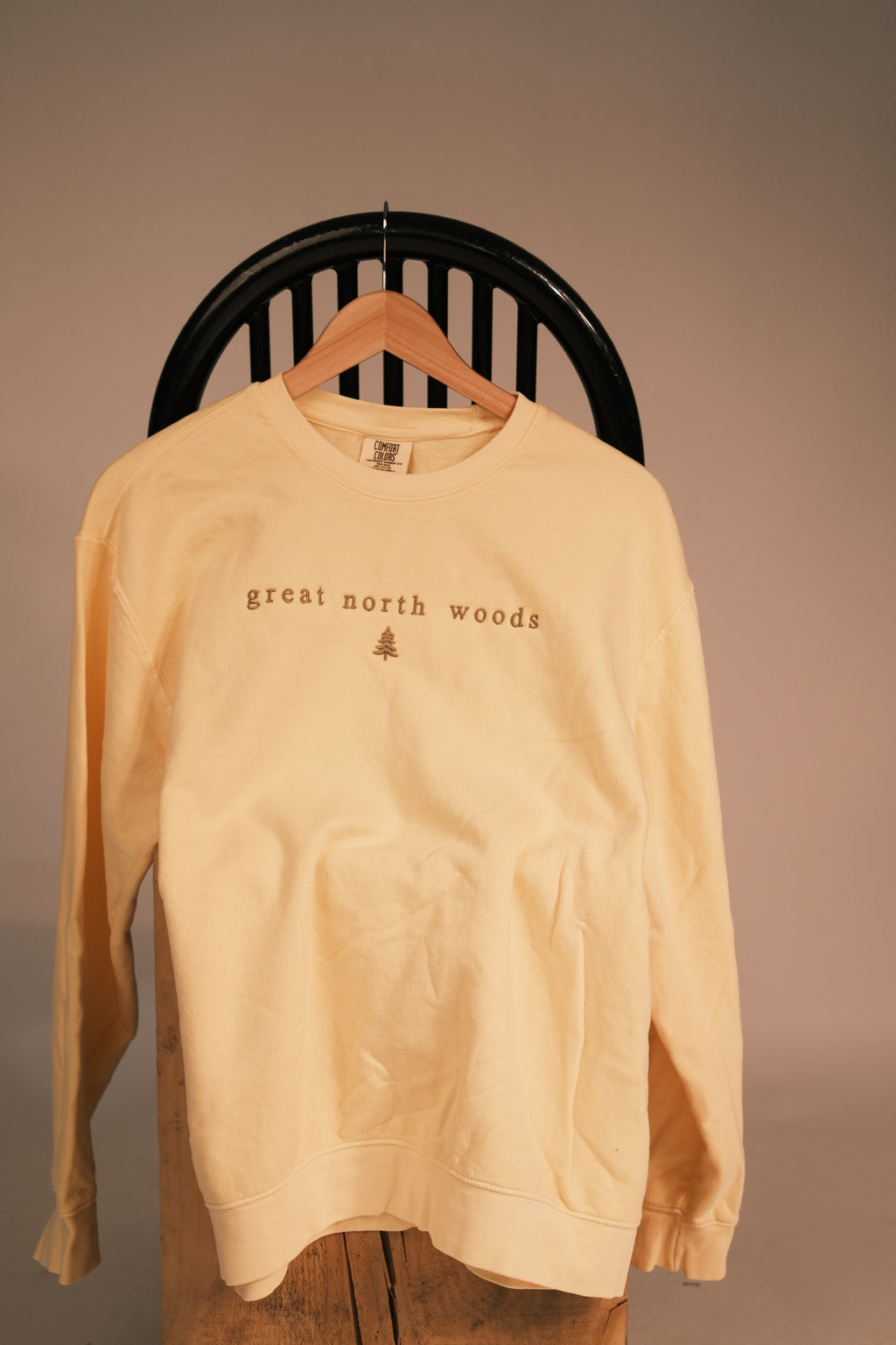 A pale yellow crewneck sweatshirt hanging on a wooden hanger from a black slatted chair, featuring the text 'great north woods' and a small tree graphic on the chest.