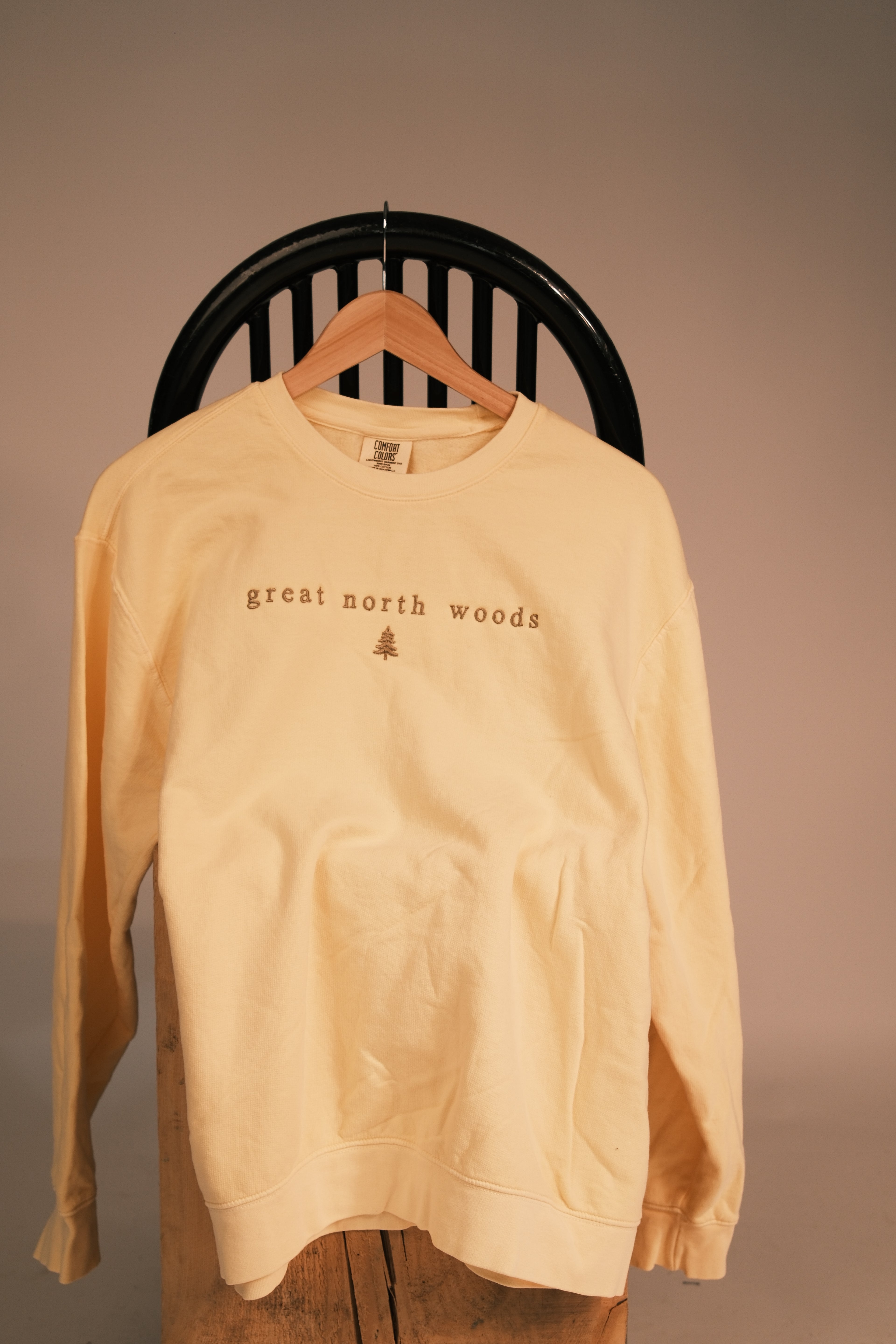 A pale yellow crewneck sweatshirt hanging on a wooden hanger from a black slatted chair, featuring the text 'great north woods' and a small tree graphic on the chest.