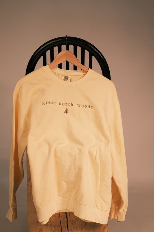 A pale yellow crewneck sweatshirt hanging on a wooden hanger from a black slatted chair, featuring the text 'great north woods' and a small tree graphic on the chest.