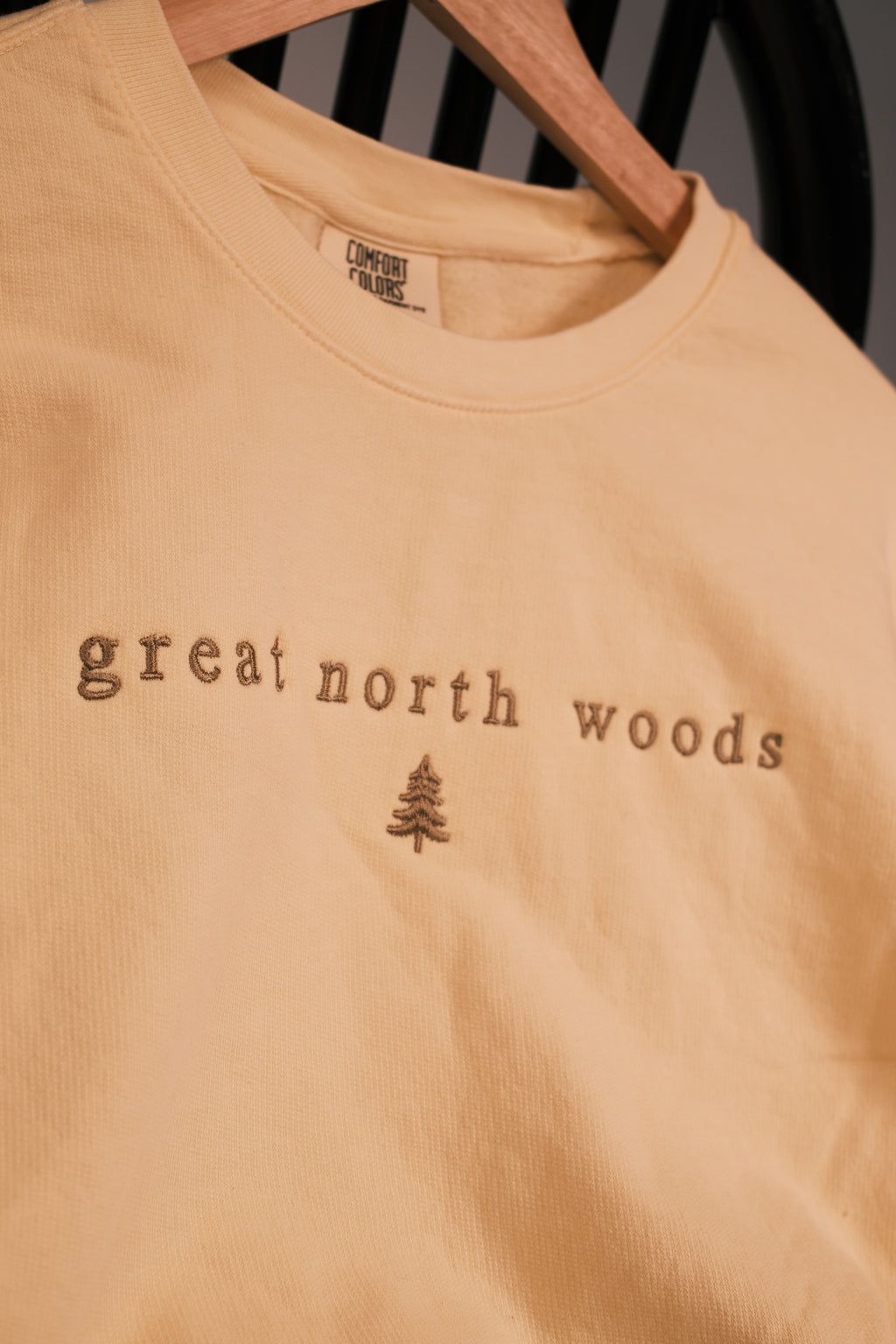A close-up of a light peach-colored crewneck t-shirt hanging on a wooden hanger. The shirt features the words 'great north woods' and a small pine tree graphic in a darker brown tone on the chest.