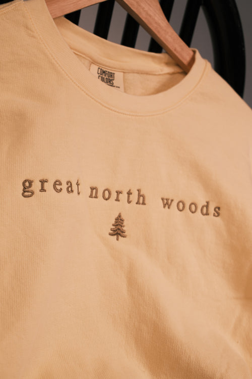A close-up of a light peach-colored crewneck t-shirt hanging on a wooden hanger. The shirt features the words 'great north woods' and a small pine tree graphic in a darker brown tone on the chest.