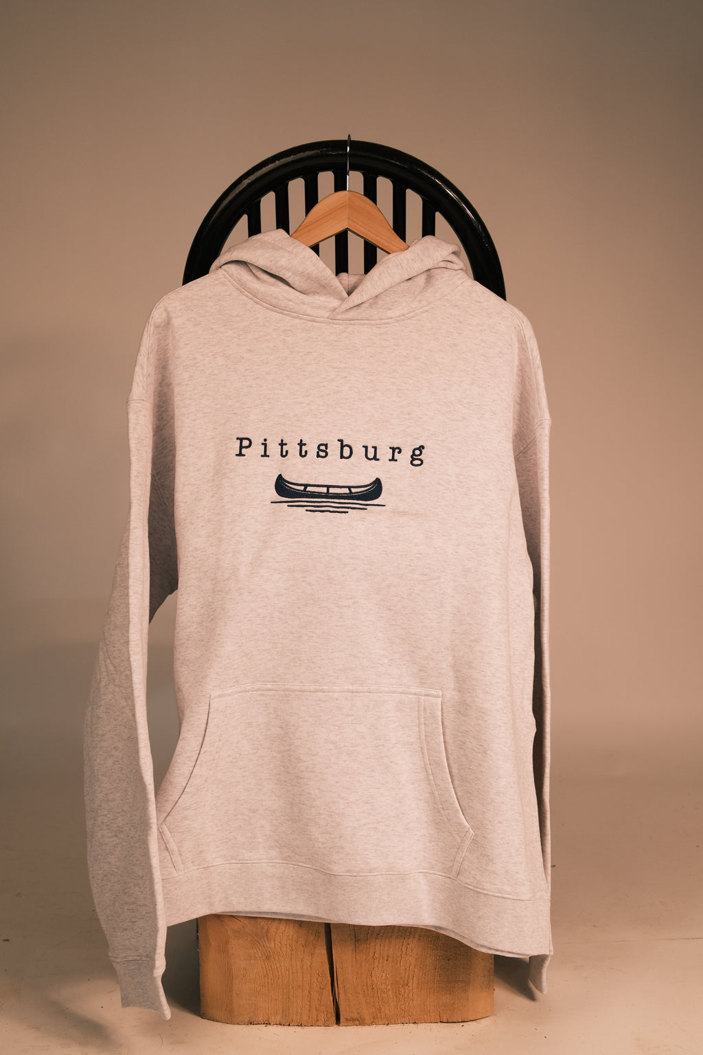A light heather gray pullover hoodie with a blue graphic print reading 'Pittsburg' and a canoe illustration, hanging on a wooden hanger against a warm beige background.