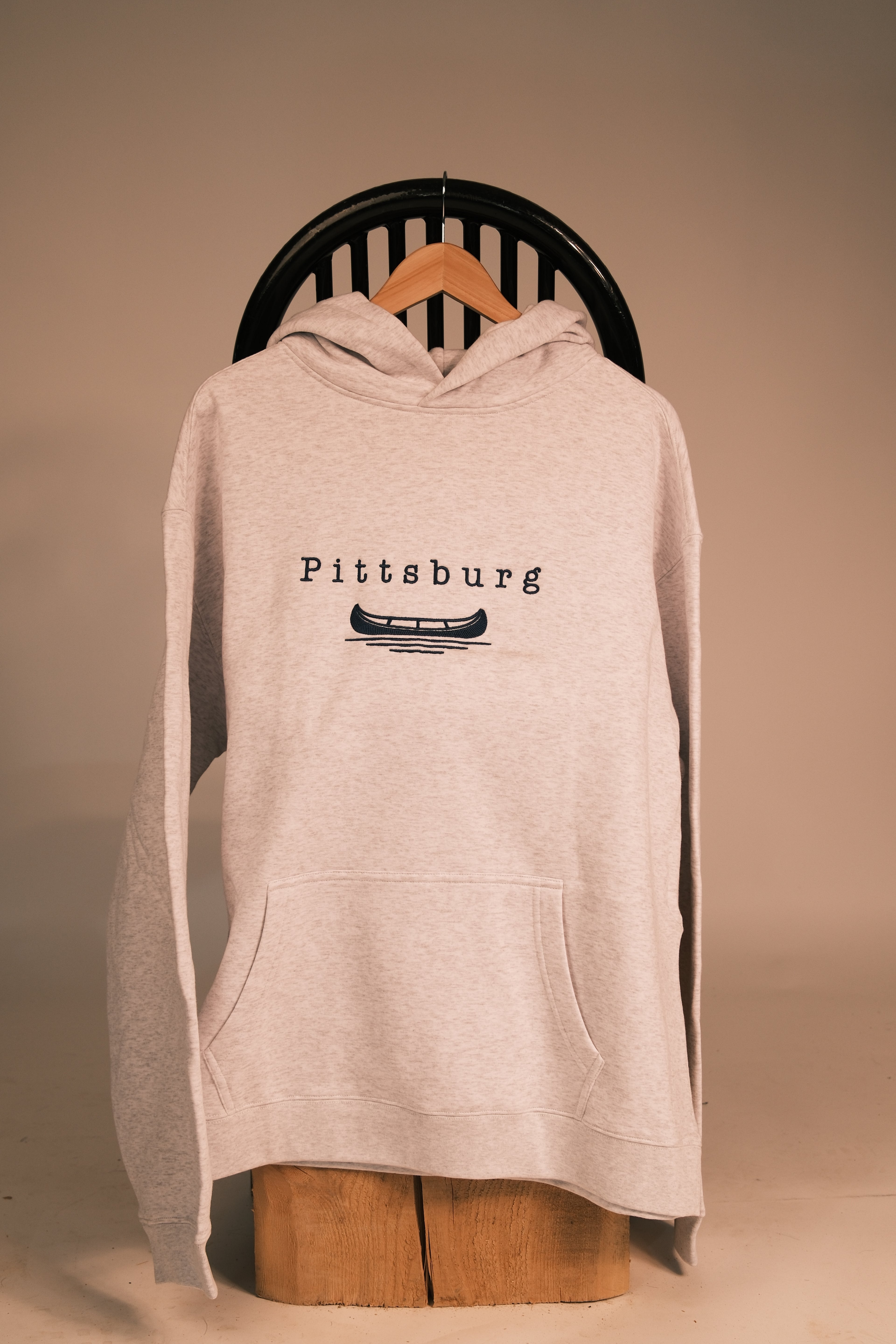 A light heather gray pullover hoodie with a blue graphic print reading 'Pittsburg' and a canoe illustration, hanging on a wooden hanger against a warm beige background.