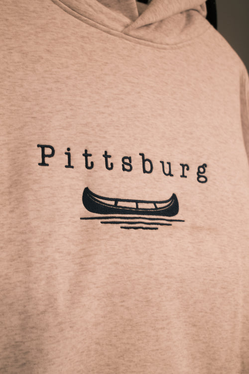 Close-up of a light heather beige hoodie with 'Pittsburg' and a canoe graphic printed in blue on the chest.