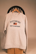 A light heather gray sweatshirt hanging on a wooden hanger from a black chair, featuring 'PITTSBURG NEW HAMPSHIRE' and an American flag graphic on the chest.