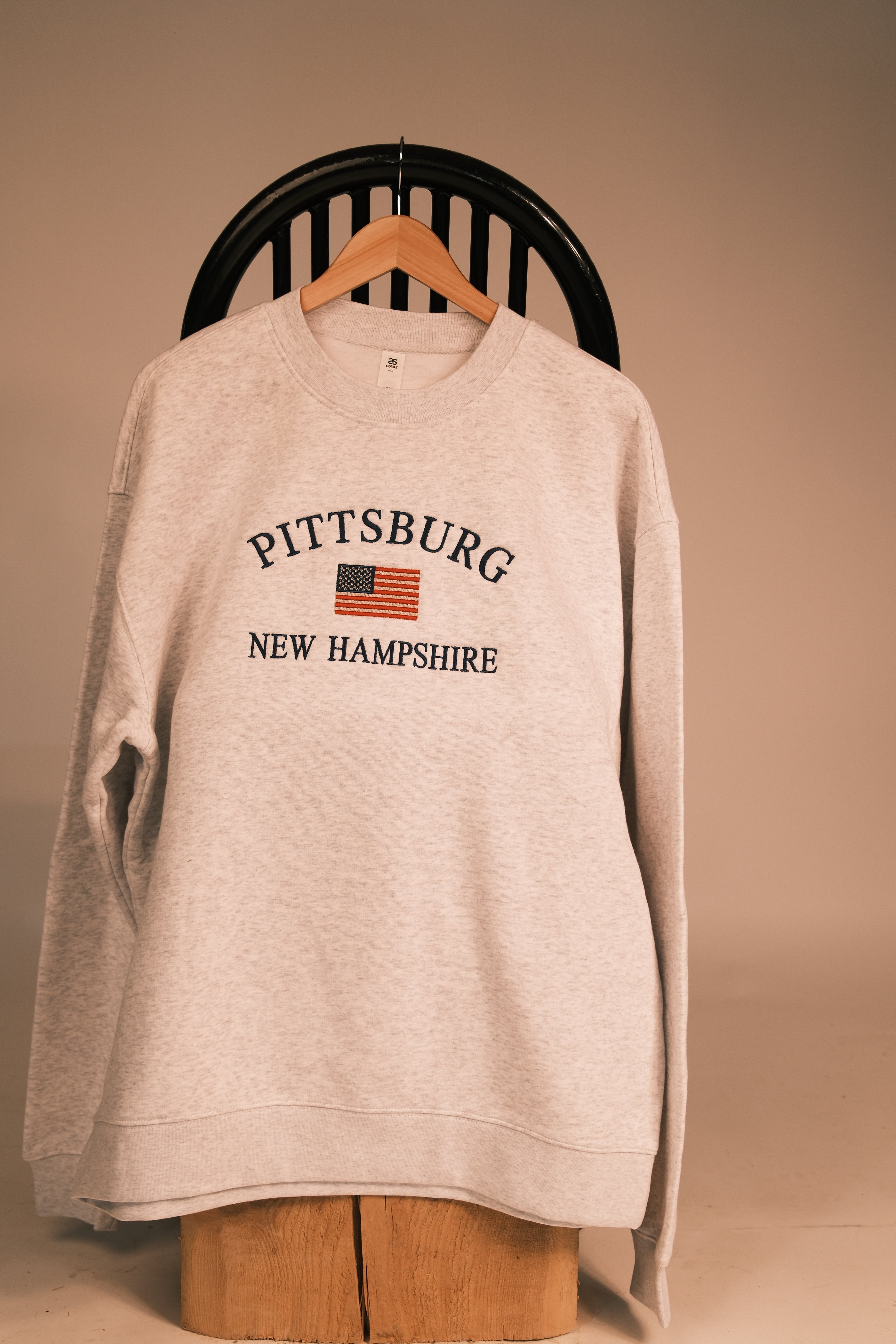 A light heather gray sweatshirt hanging on a wooden hanger from a black chair, featuring 'PITTSBURG NEW HAMPSHIRE' and an American flag graphic on the chest.