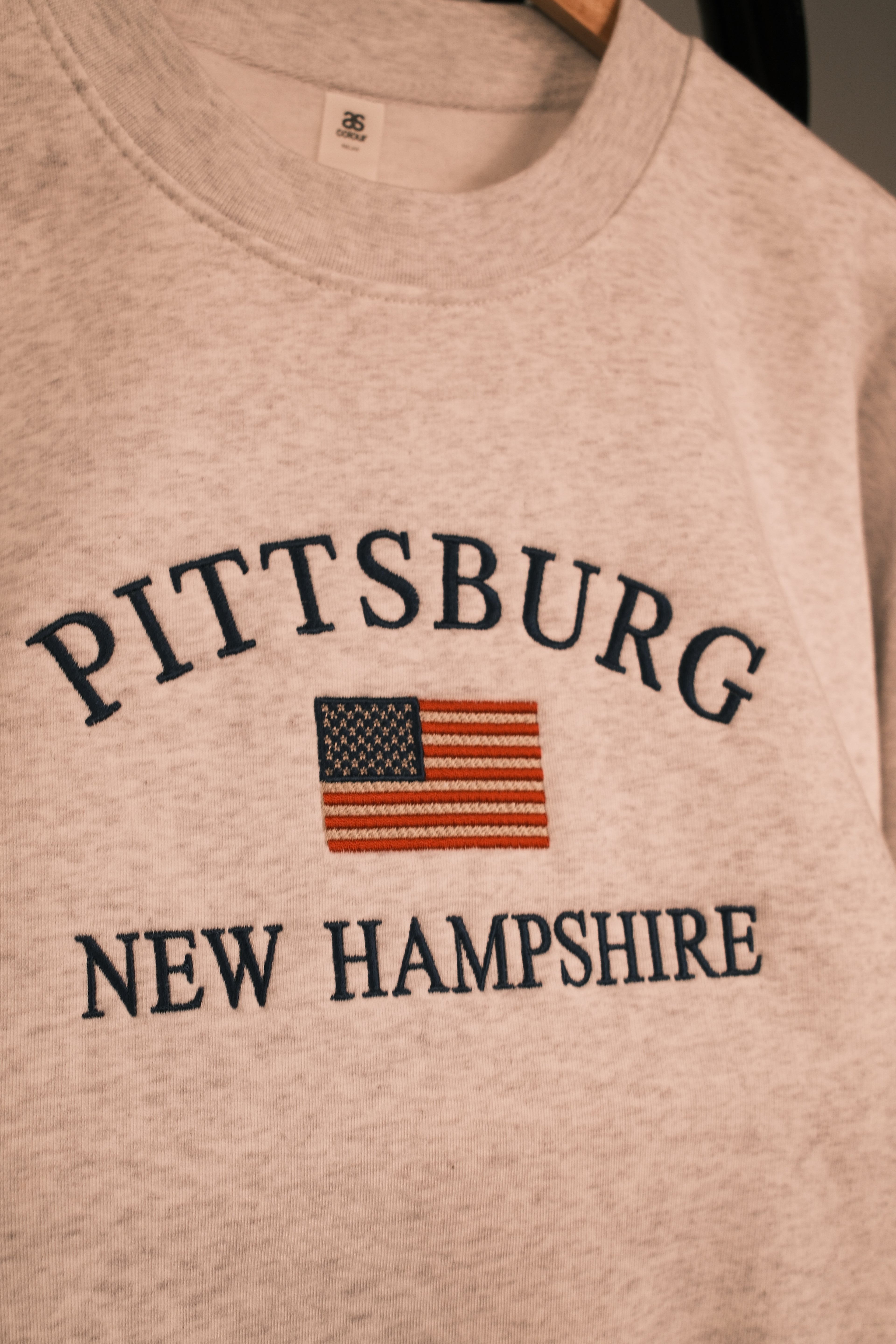 Close-up of a light heather gray crewneck sweatshirt hanging on a wooden hanger, featuring 'PITTSBURG' and 'NEW HAMPSHIRE' in dark navy text with an embroidered American flag graphic in the center.
