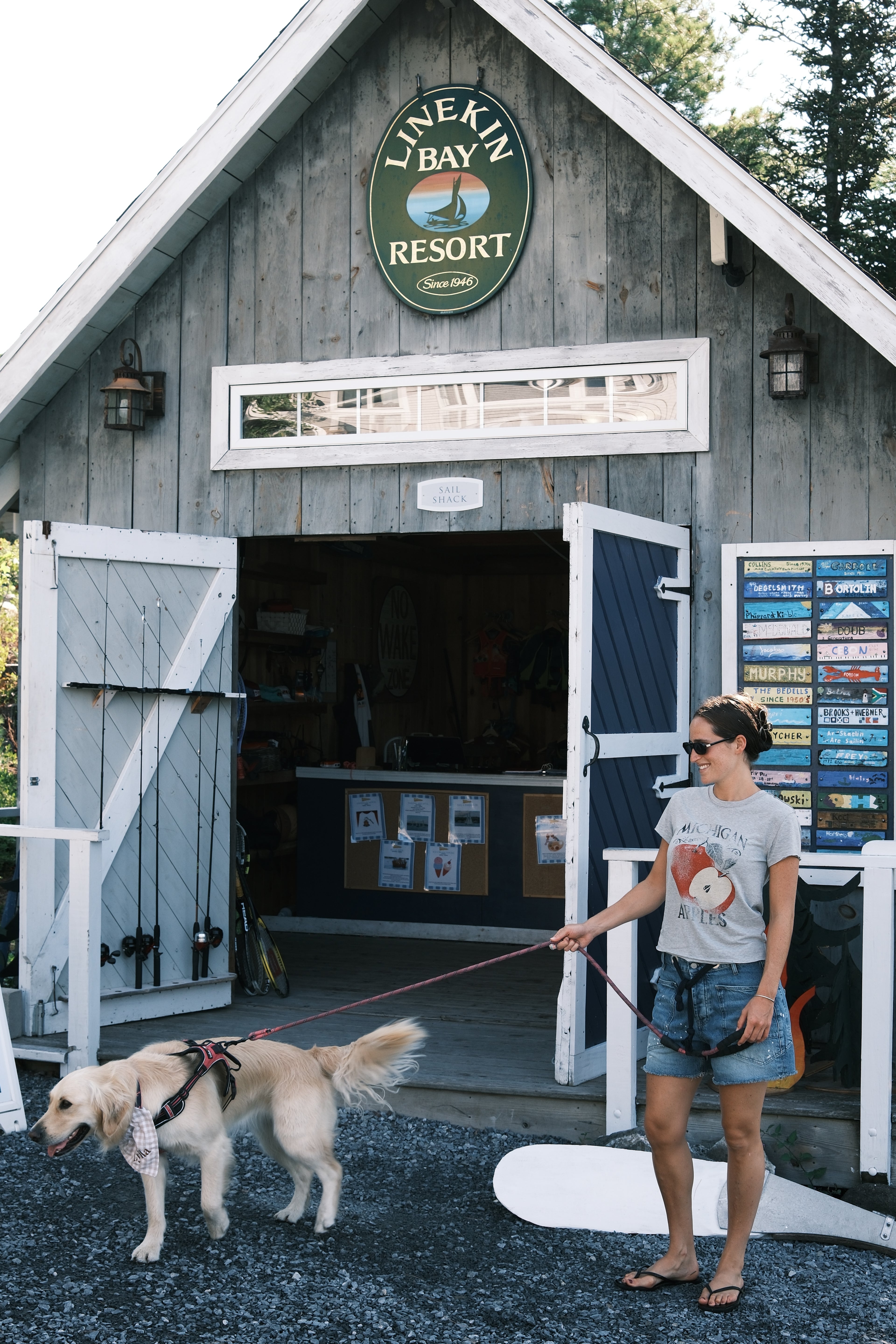 A woman stands on a gravel driveway in front of a rustic gray wooden resort building, holding a leash attached to a light-colored dog. A large circular sign above the entrance reads 'Lonekin Bay Resort'.