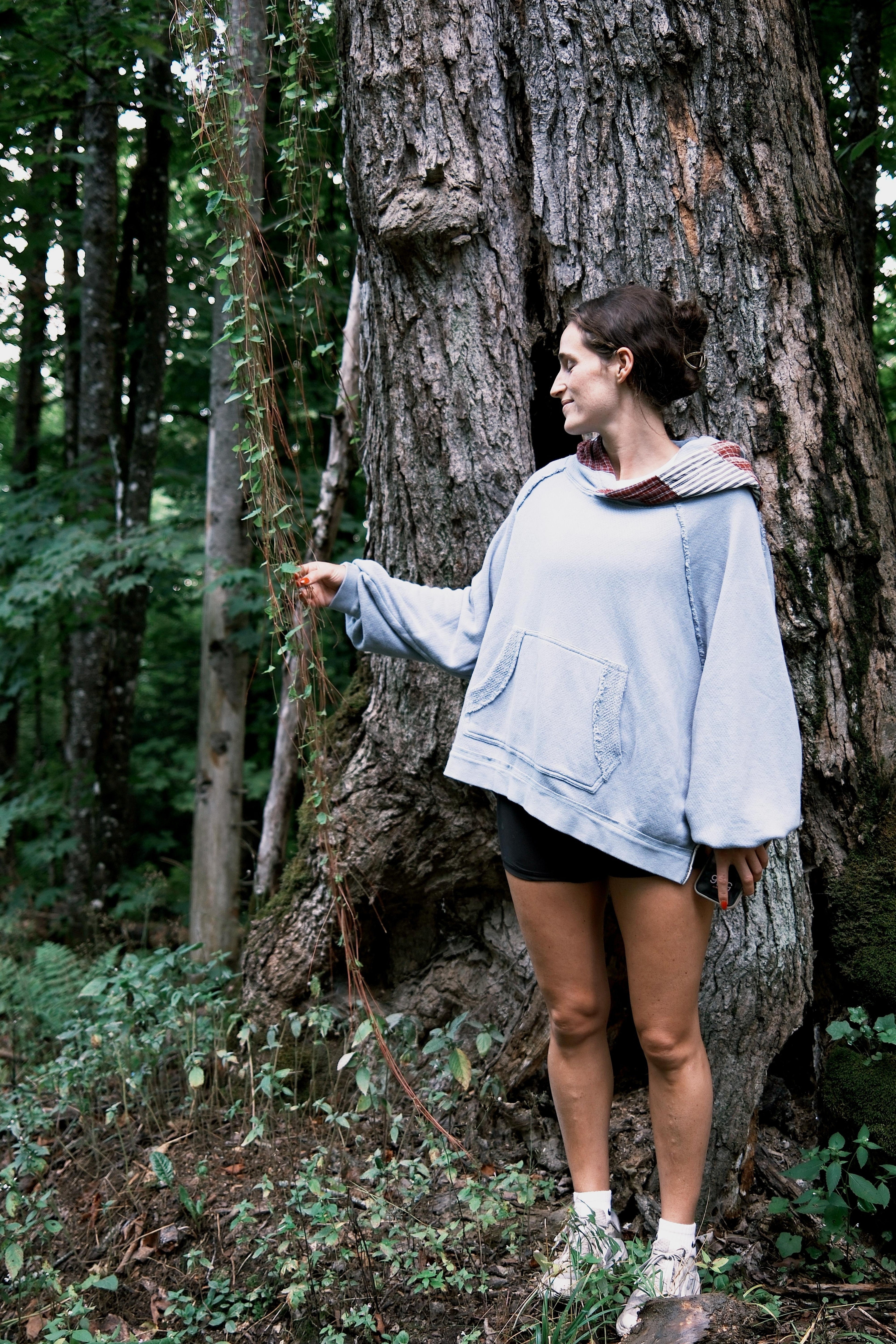 A woman wearing a light blue oversized denim jacket, black shorts, and white socks lies on her back on a large tree trunk in a forest.