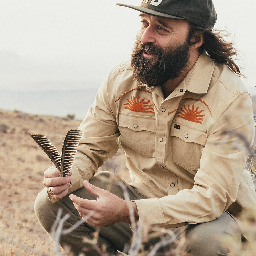 Man in beige shirt with orange designs, holding a comb, outdoors.
