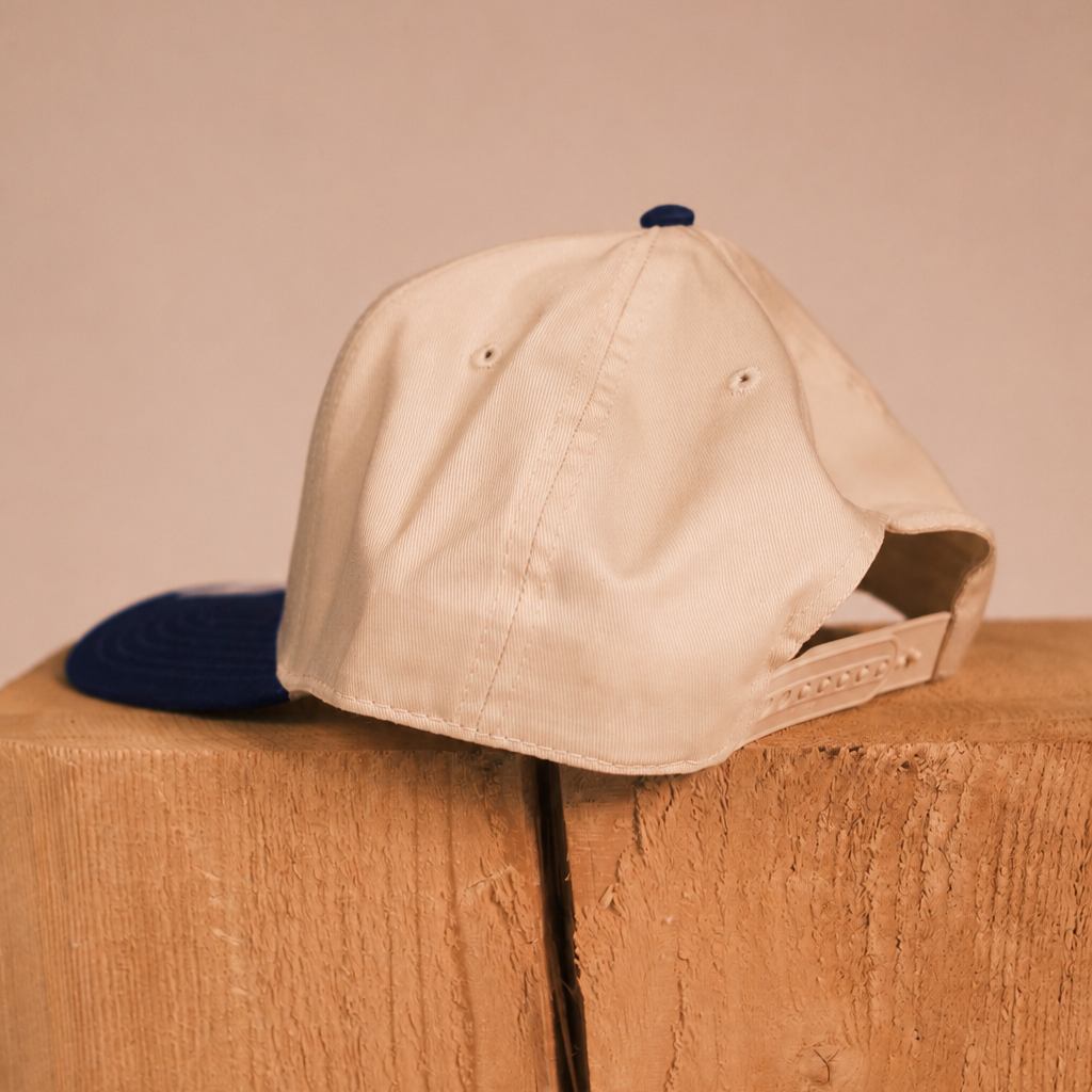 A beige baseball cap with a navy blue brim and matching navy button on top, resting on a rustic wooden block against a solid beige background.