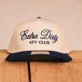 A cream-colored snapback cap with a navy blue brim, featuring 'Extra Dirty ATV CLUB' embroidered on the front. The cap is displayed on a rustic wooden block against a neutral beige background.