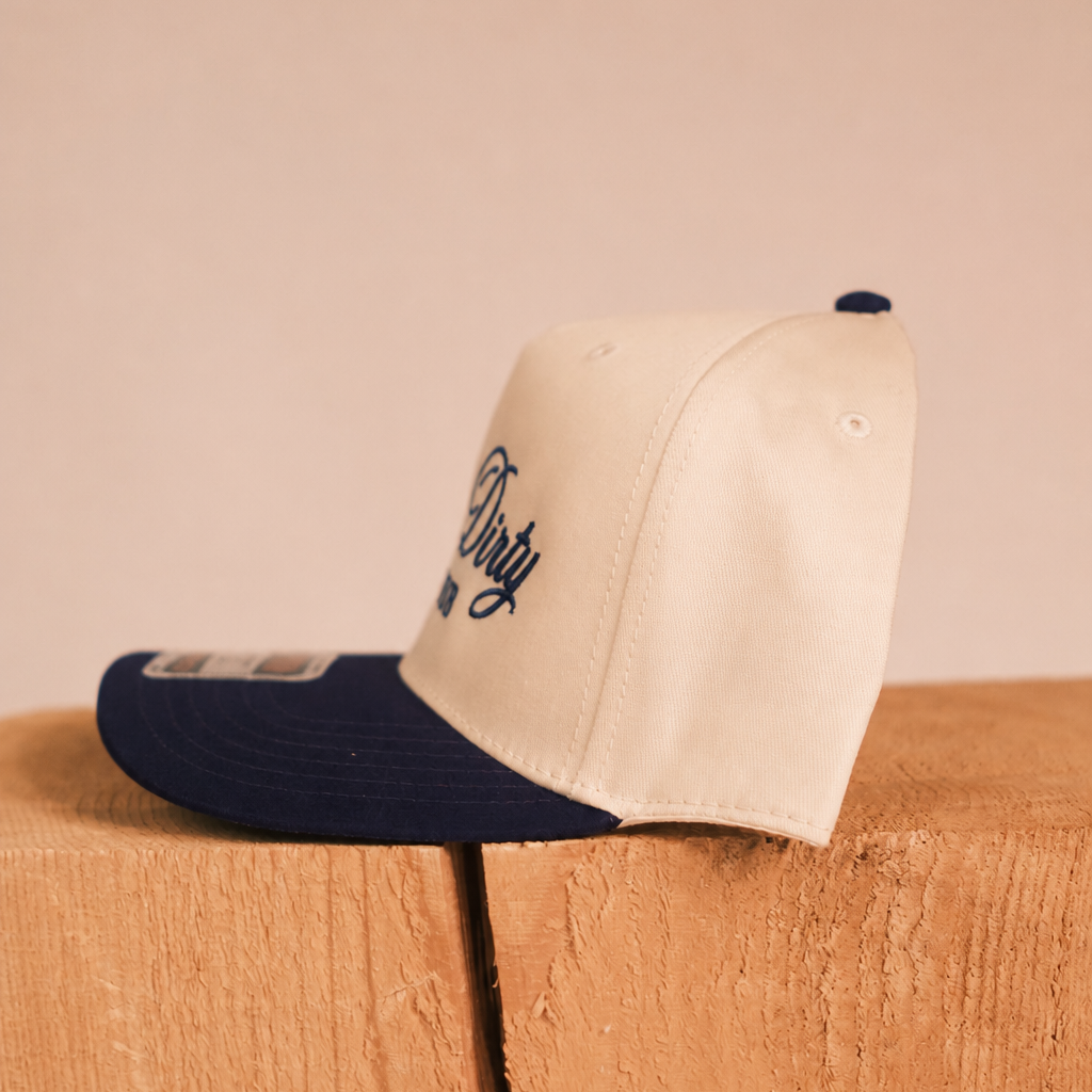 A cream and navy blue trucker-style cap with embroidered script on the front, resting on a wooden block against a solid beige background.