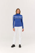 Woman wearing a blue patterned sweater with white pants on a light gray background