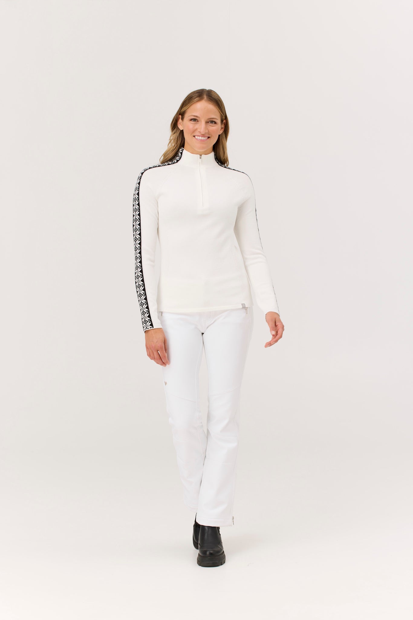 Woman wearing a white turtleneck with black accents and white pants on a light gray background
