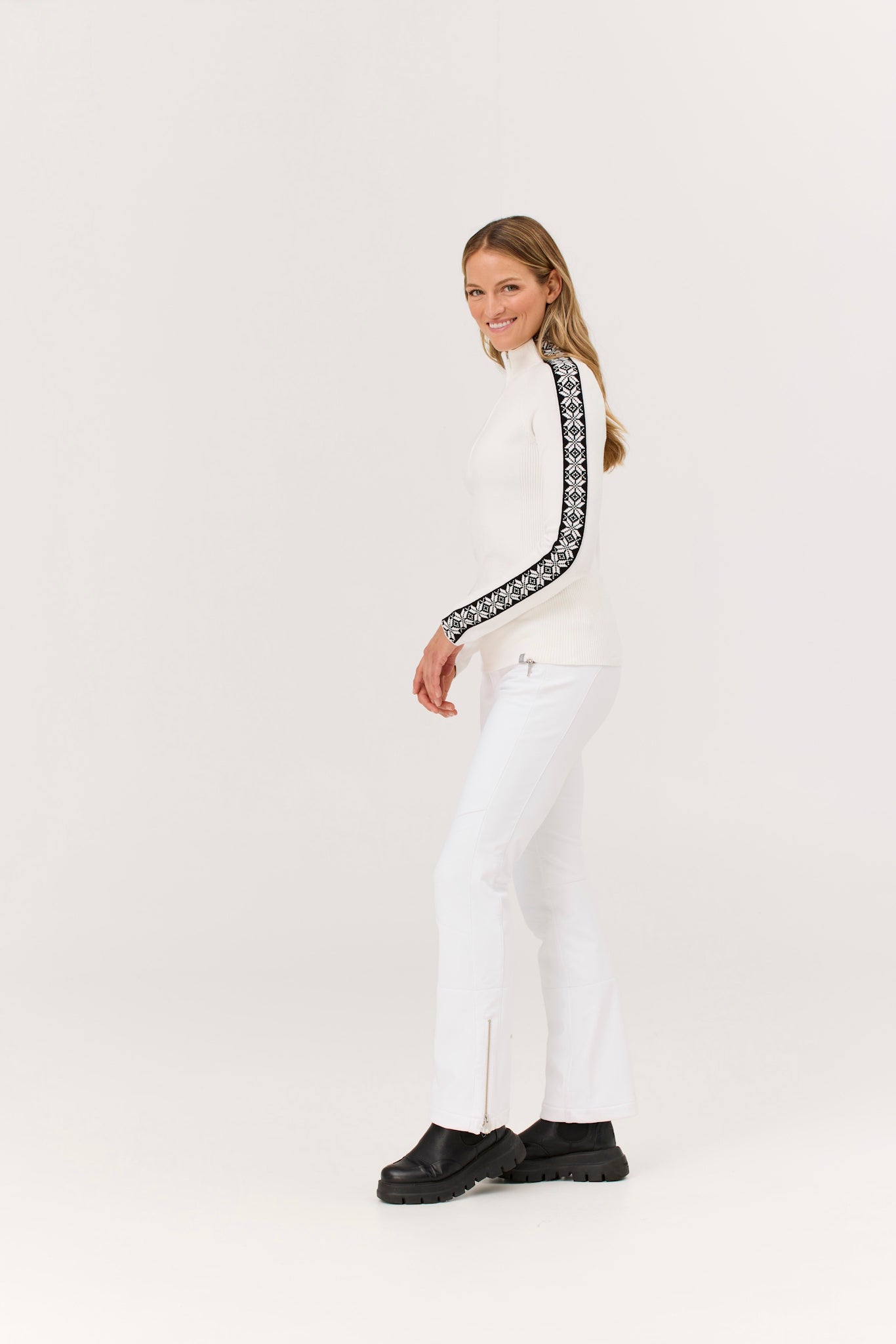 Woman wearing a white turtleneck with black patterned trim and white pants on a white background